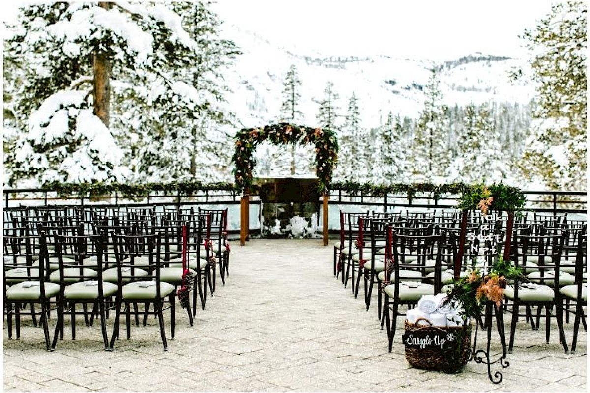 Snowy outdoor wedding setting with chairs, floral arch, and mountainous background, surrounded by snow-covered trees and serene winter scenery.