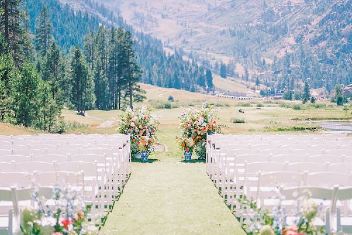 An outdoor wedding setup with rows of white chairs on either side of a grassy aisle, decorated with flowers, mountains, and trees in the background.