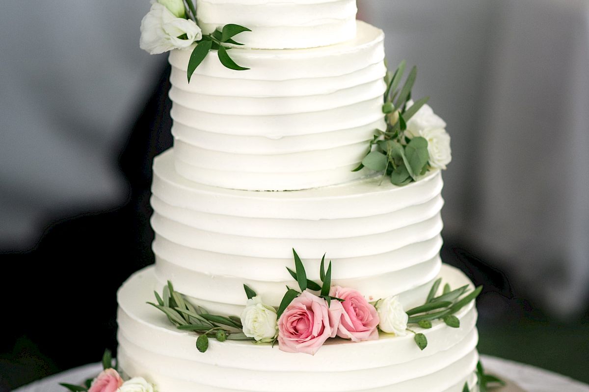 A three-tiered white wedding cake adorned with pink and white flowers and green leaves, placed on a table with plates and a gold cake server.