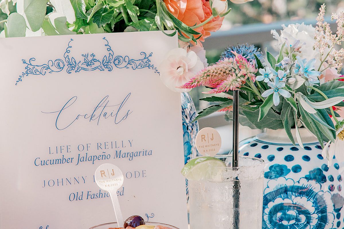 The image shows two cocktails, a floral arrangement, a decorated vase, and a menu card with cocktail names on a table outdoors, potentially at an event.