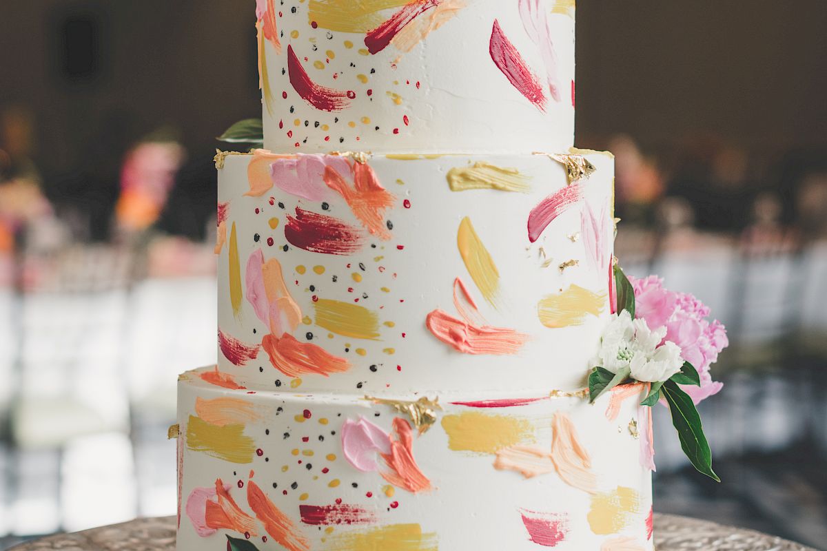 A three-tiered cake with colorful brushstroke decorations and floral accents on top and around, placed on an ornate silver cake stand.