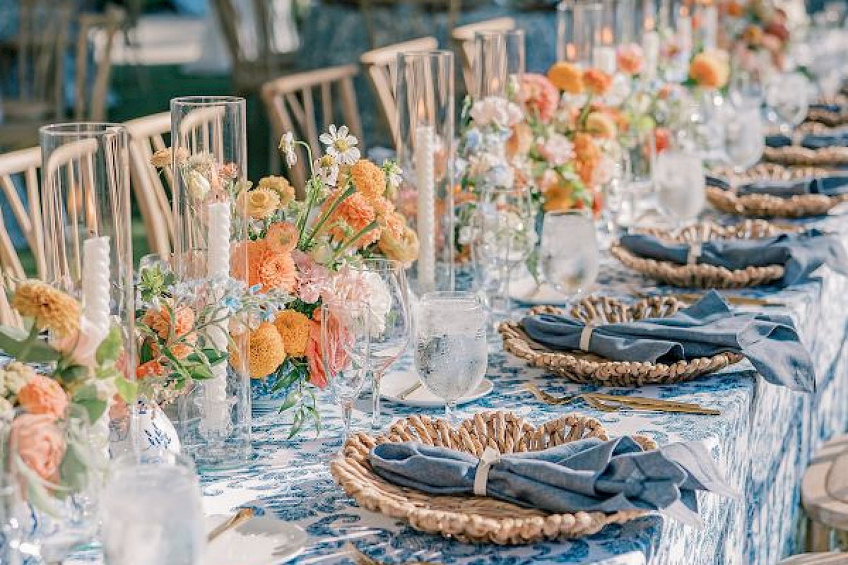 A long, elegantly set outdoor table with floral arrangements, candles, woven placemats, and blue napkins is ready for a gathering or celebration.