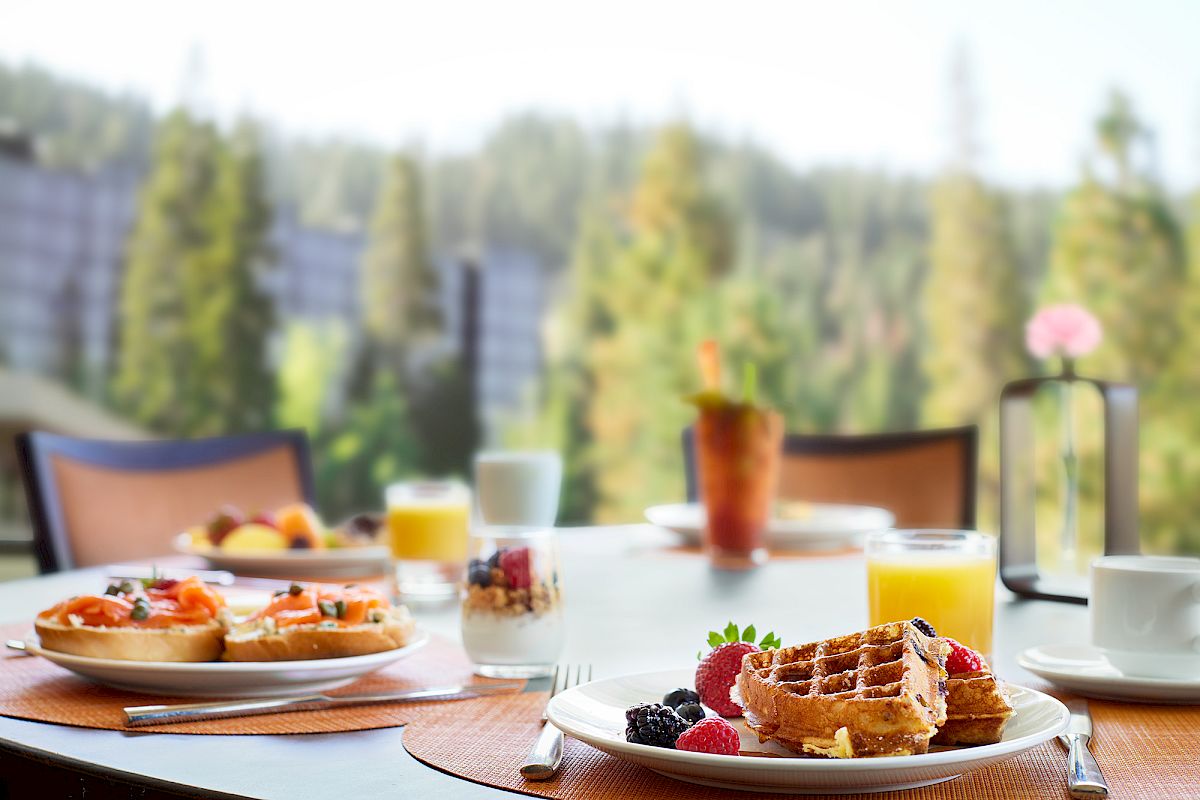 A breakfast table with waffles, juice, fruit, and a parfait, set against a blurry forest view.