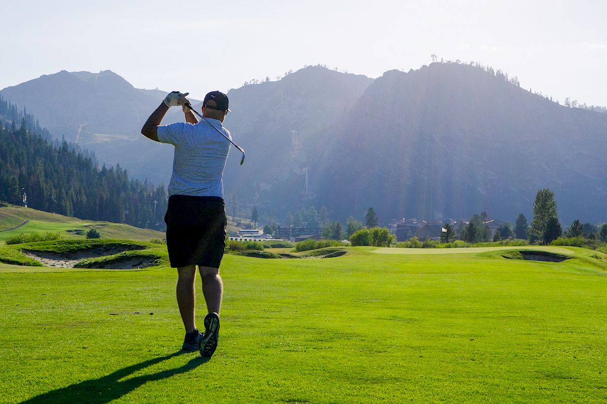 A person is playing golf on a grassy golf course surrounded by mountains and trees under a clear sky with the sun shining brightly.