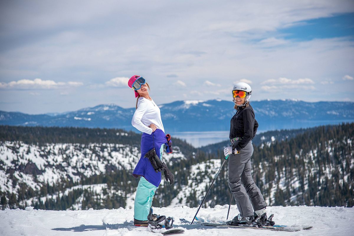 Two people in ski gear posing on a snow-covered mountain with scenic views of trees and mountains in the background.