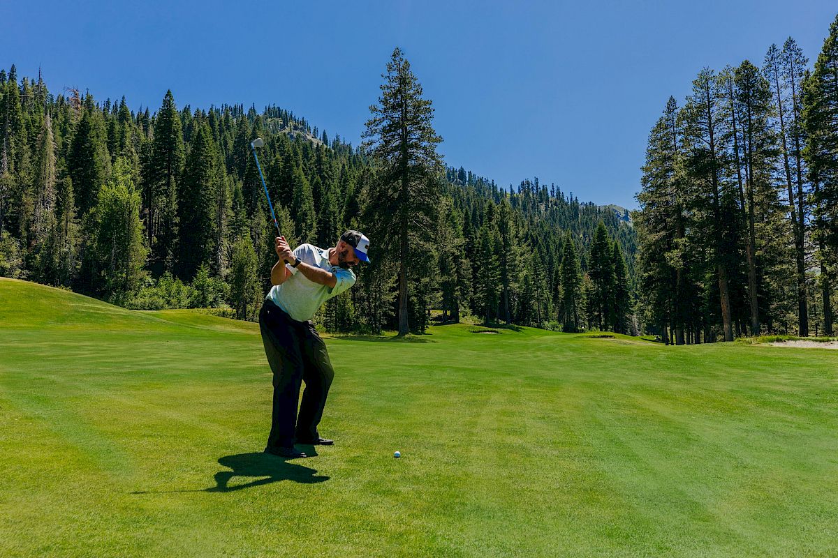 A golfer is mid-swing on a lush, green golf course surrounded by tall trees and mountains under a clear blue sky.