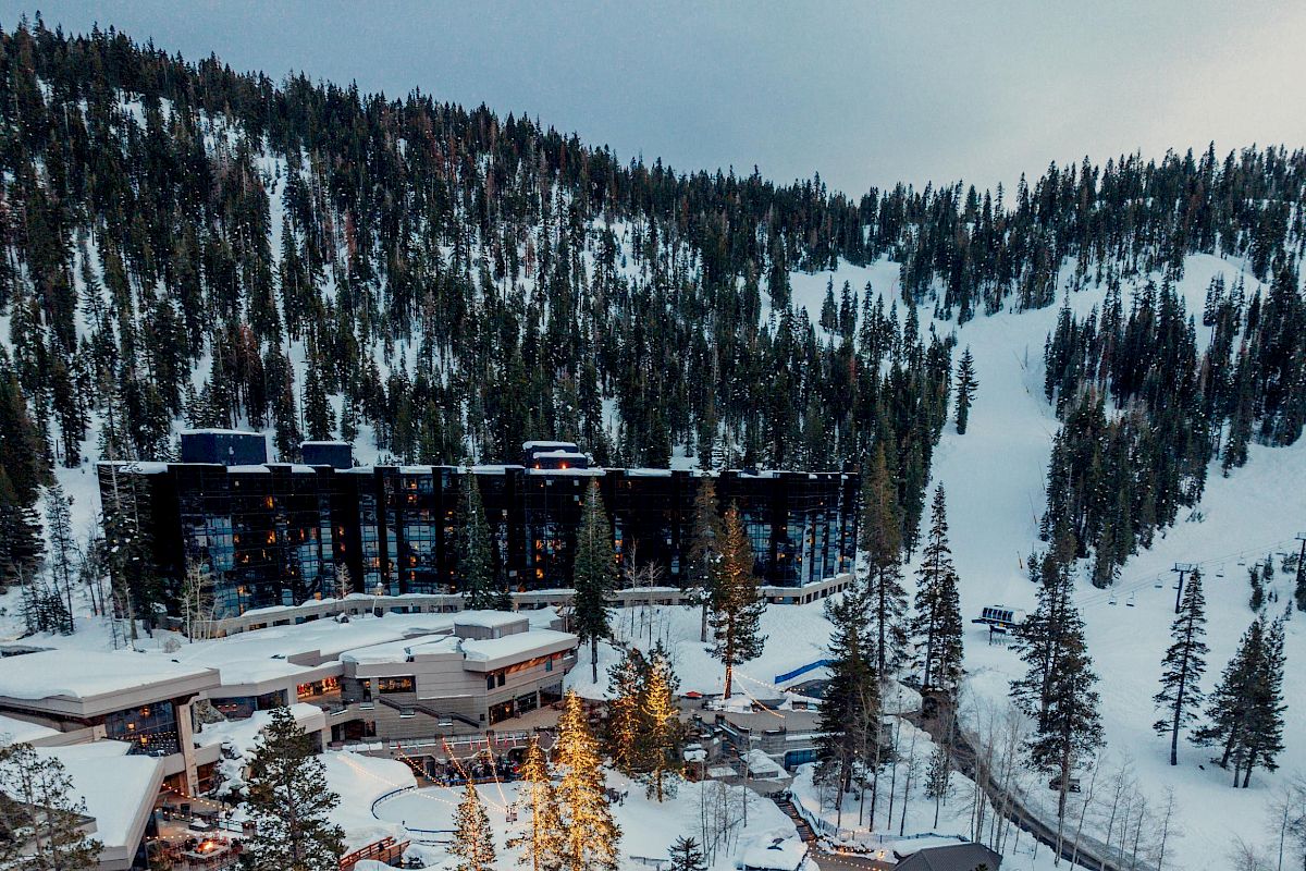 A snowy mountain resort with outdoor pools, surrounded by dense evergreen trees. The buildings are nestled within the snowy landscape and forest.