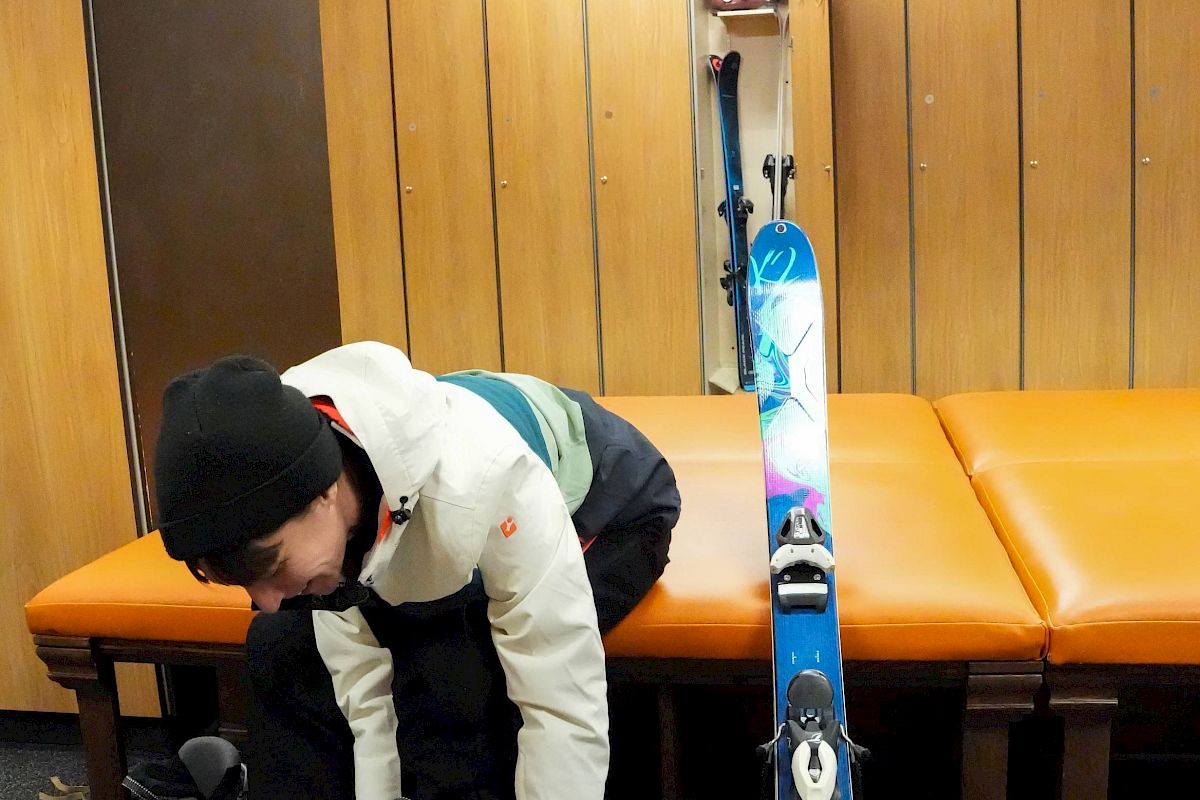 A person is sitting on an orange bench, adjusting their ski boot with a colorful ski leaning against the bench in a wooden room.