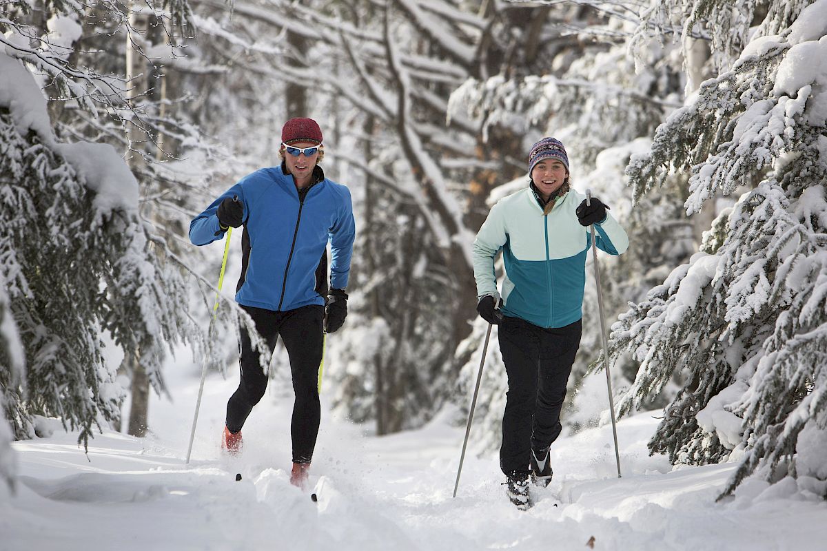 Two people cross-country skiing on a snow-covered trail in a forest, dressed in winter gear and smiling.