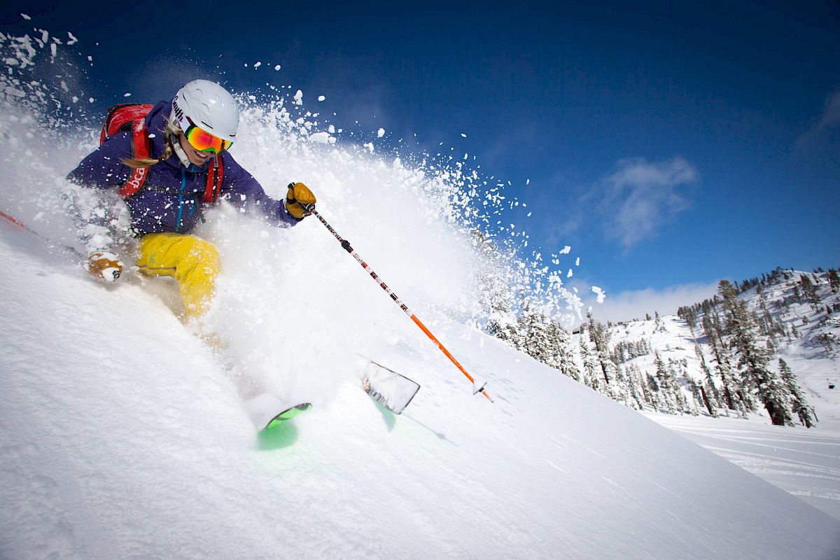 A person is skiing down a snowy slope, creating a spray of powder snow, with a clear blue sky and a mountainous backdrop in the distance.