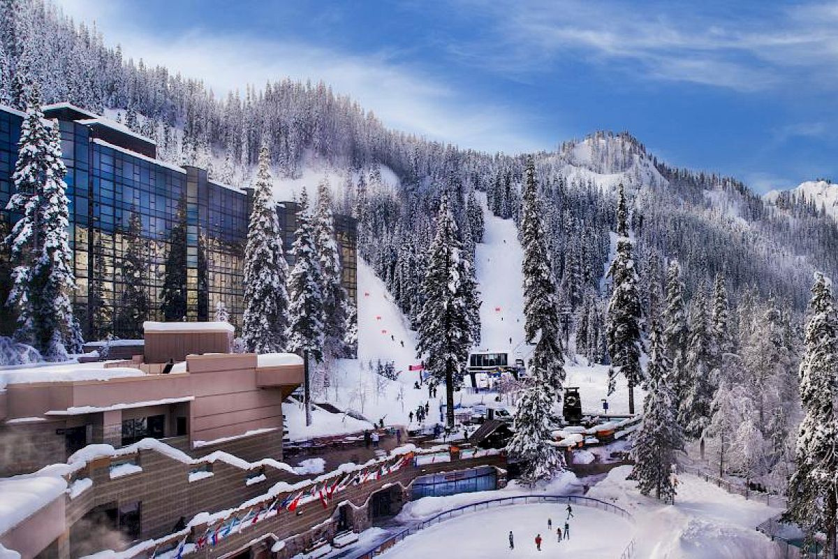 A beautiful winter scene with snowy mountains, a ski slope, a lodge, and people skating on a frozen pond surrounded by trees and buildings.