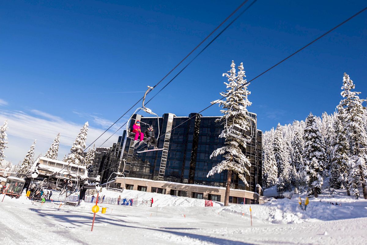 The image shows a snowy ski resort with people on a ski lift, a large building, and snow-covered trees under a clear blue sky.