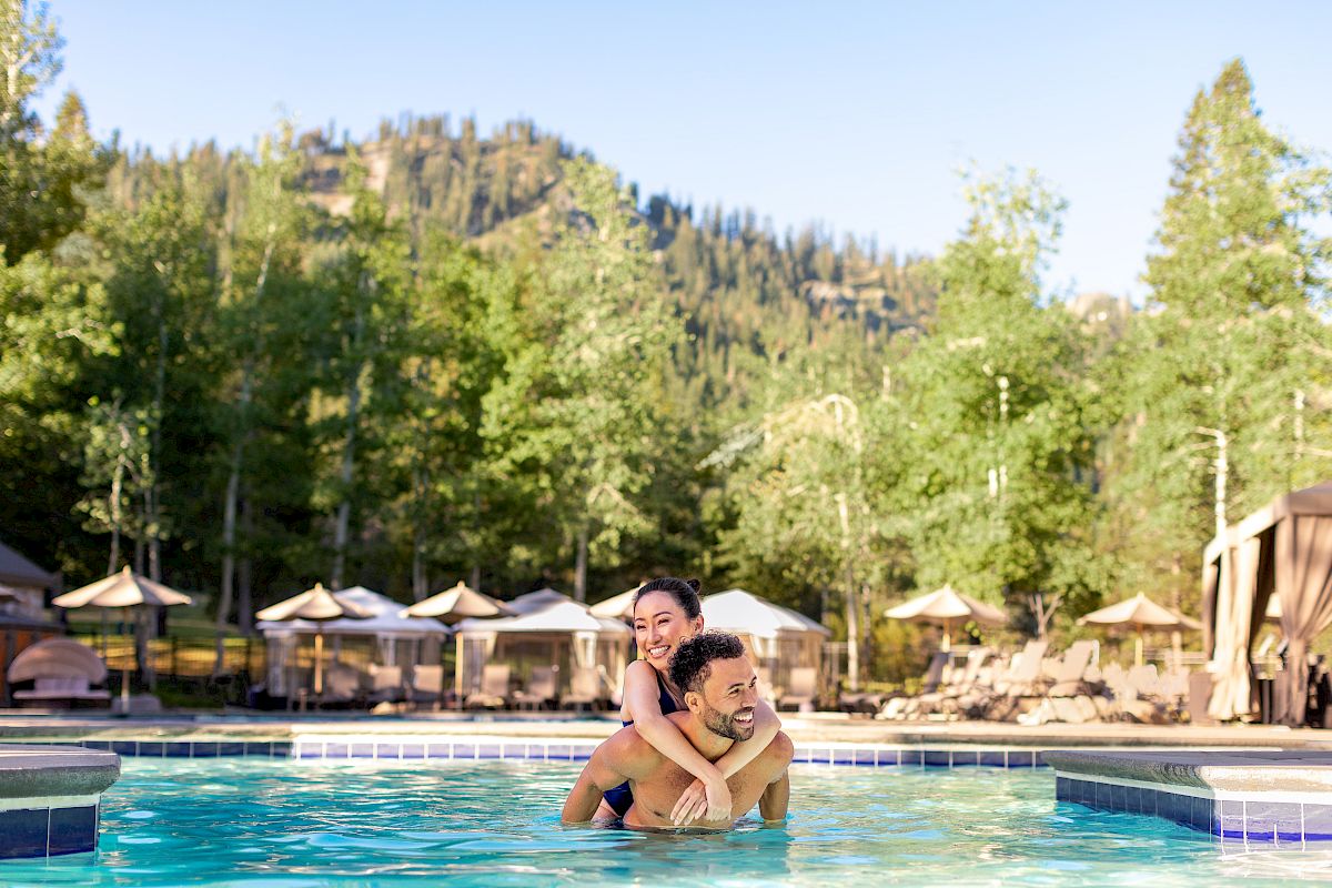 A couple is enjoying a playful moment in a pool, set against a backdrop of trees and cabanas with a mountainous landscape in the background.