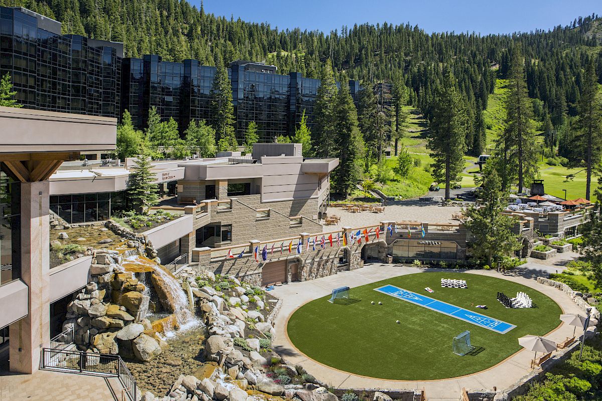 The image shows a modern resort nestled in a forested mountainous area, featuring buildings, a fountain, and a turf area with flags and seating.
