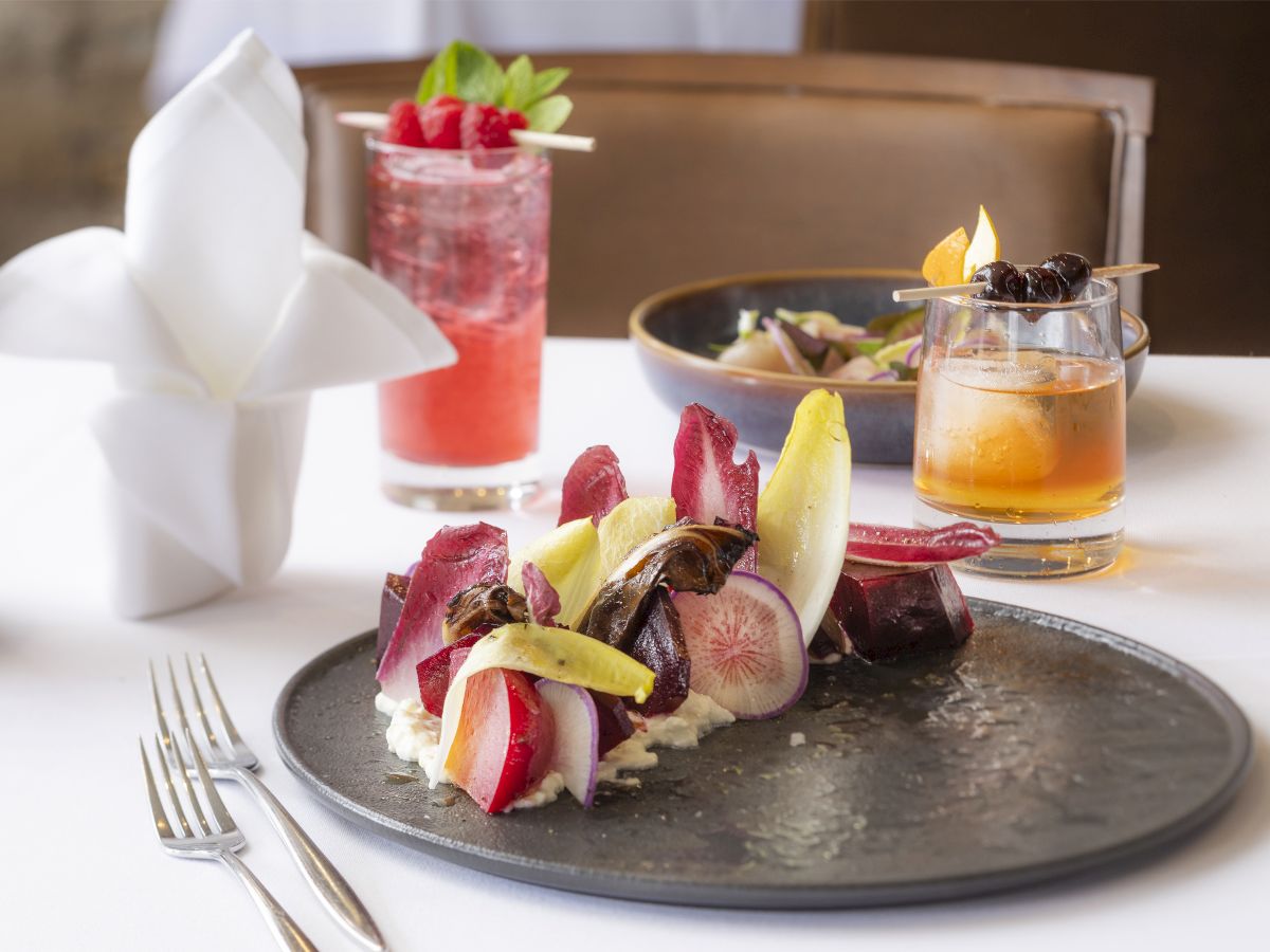 A stylishly arranged plated dish featuring vegetables, alongside two vibrant cocktails and a decorative napkin on a white tablecloth.