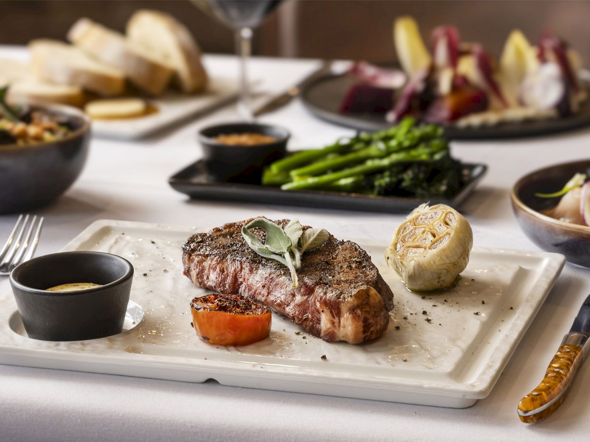 A steak plate with garnish, grilled tomato, and sauce, alongside sides of bread, broccolini, mixed vegetables, and wine are on a table.