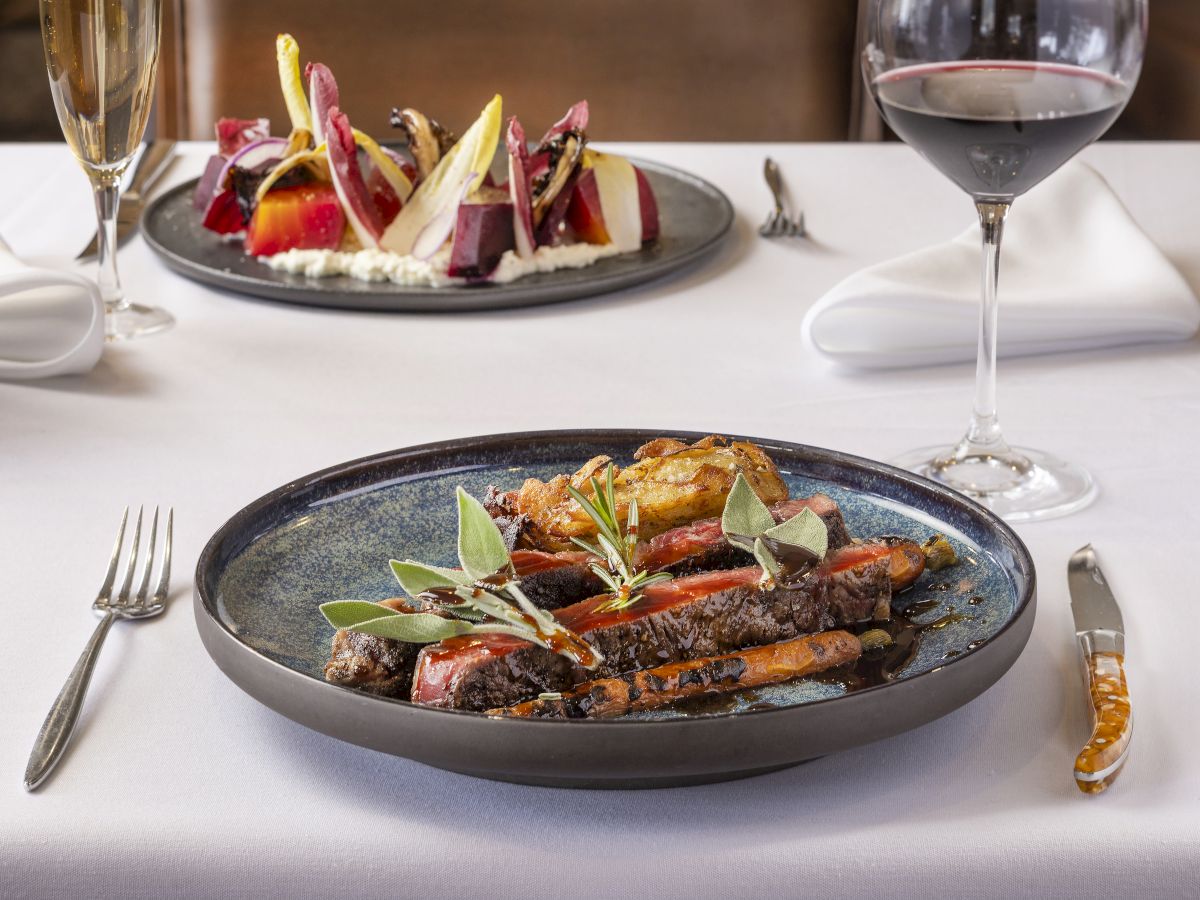 The image shows a fine dining setup with a plate of grilled vegetables, another dish in the background, and a glass of red wine on a white tablecloth.