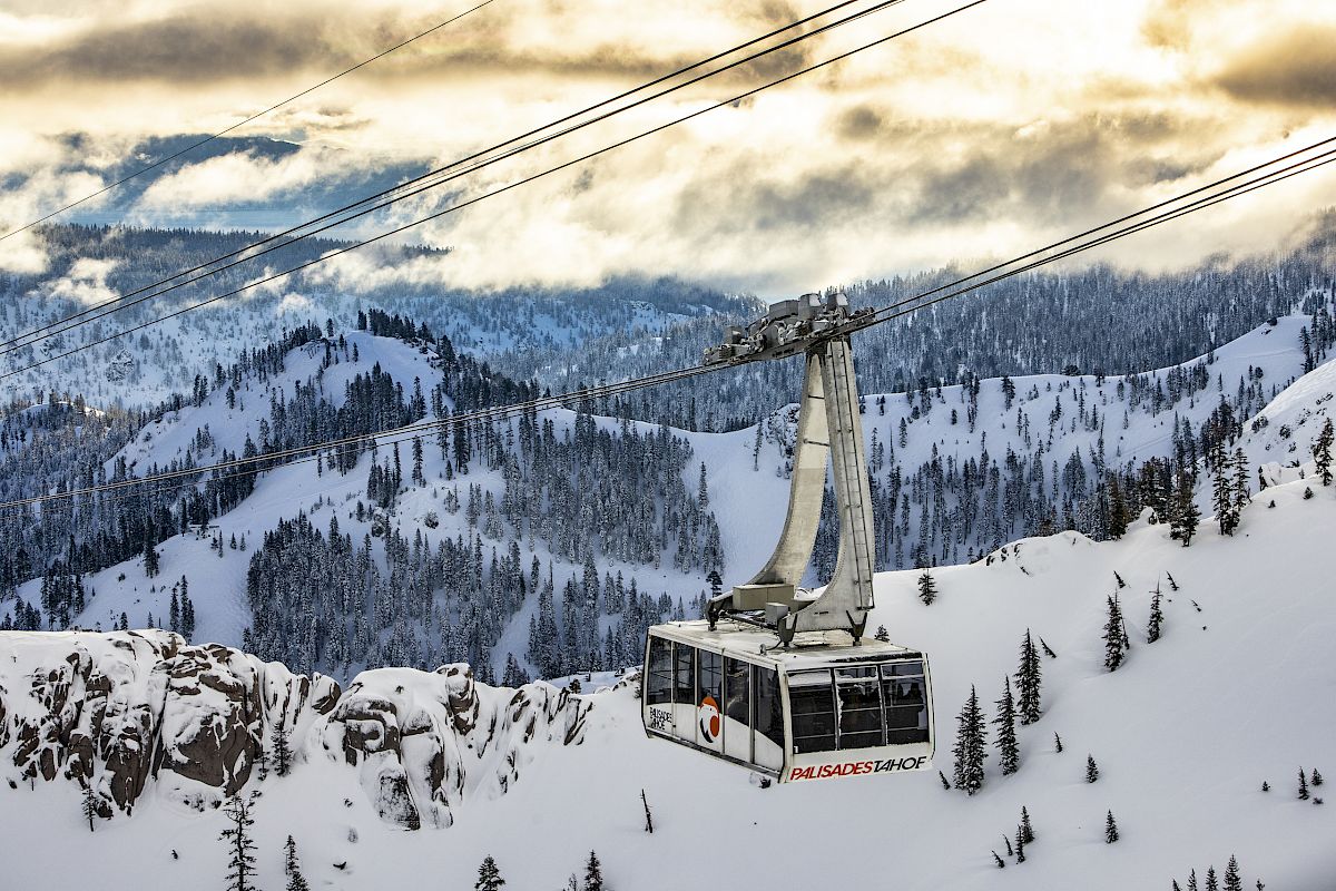A cable car transports passengers over a snowy mountain landscape, surrounded by trees under a dramatic cloudy sky.