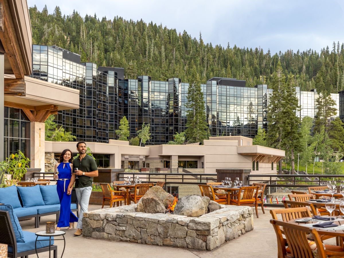 A couple stands by an outdoor fire pit surrounded by seating, with a modern glass building and wooded hillside in the background.