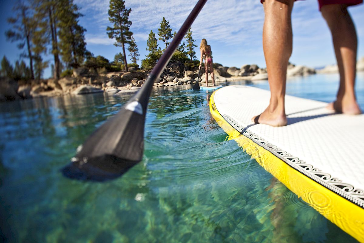 A person is paddleboarding on clear, turquoise water with trees and rocks in the background. Another person stands onshore in the distance.