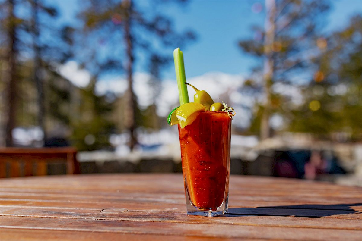 A Bloody Mary cocktail garnished with celery, lemon, and olives sits on a wooden table outdoors, surrounded by a blurred forest background.