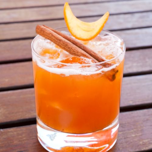 A glass of an orange-colored cocktail topped with a cinnamon stick and a slice of citrus peel, placed on a wooden table.