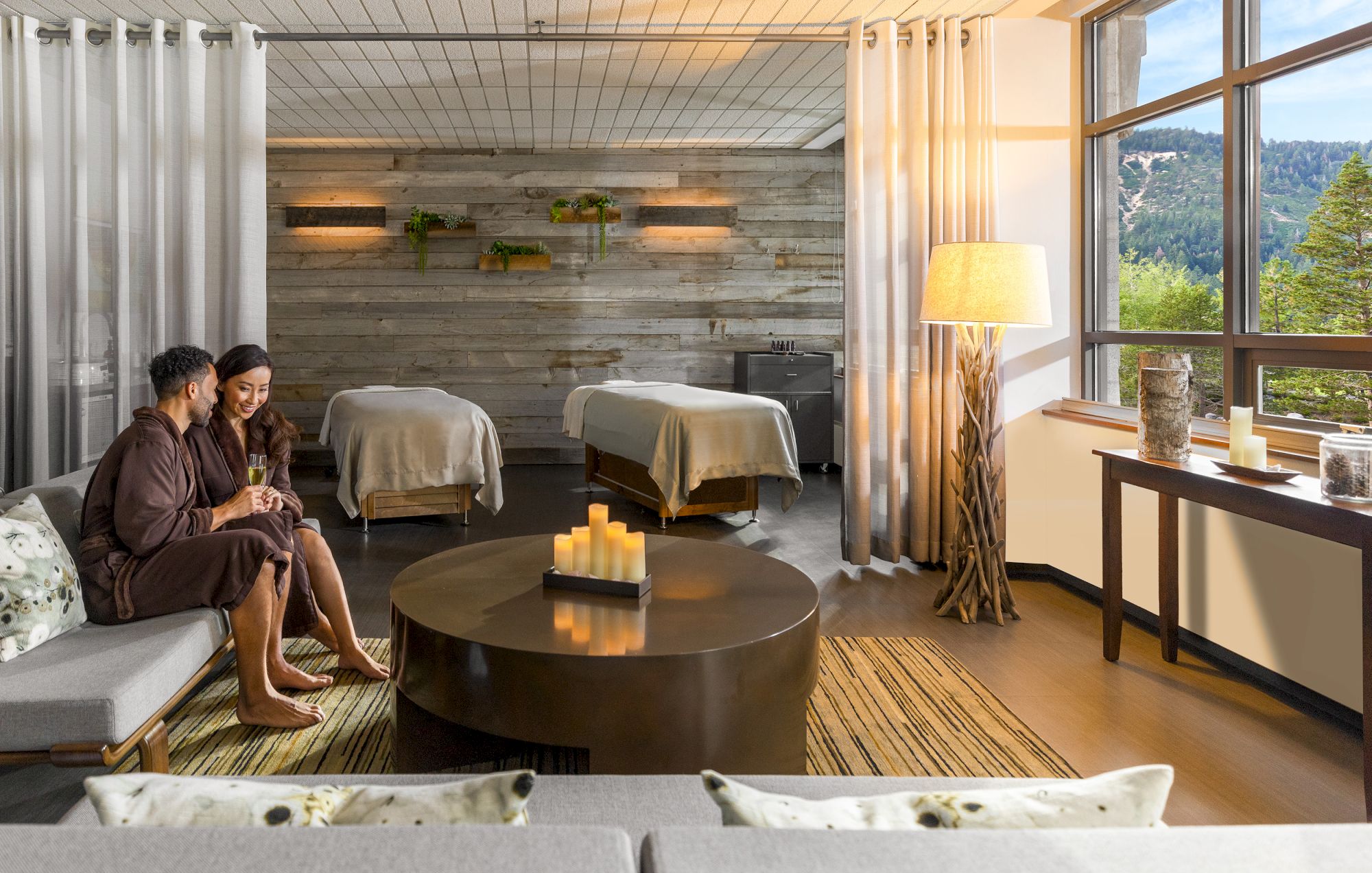 Two people in a spa lounge with massage tables, comfortable seating, and a view of the outdoors through large windows.